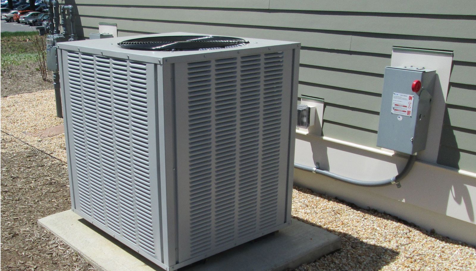 A residential outdoor unit on a concrete pad beside a house with green siding, with its electrical disconnect on the wall